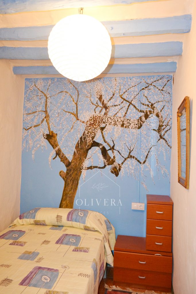 Habitación 1 con cama doble, armario y mesita. Mural de un almendro pintado en la pared.